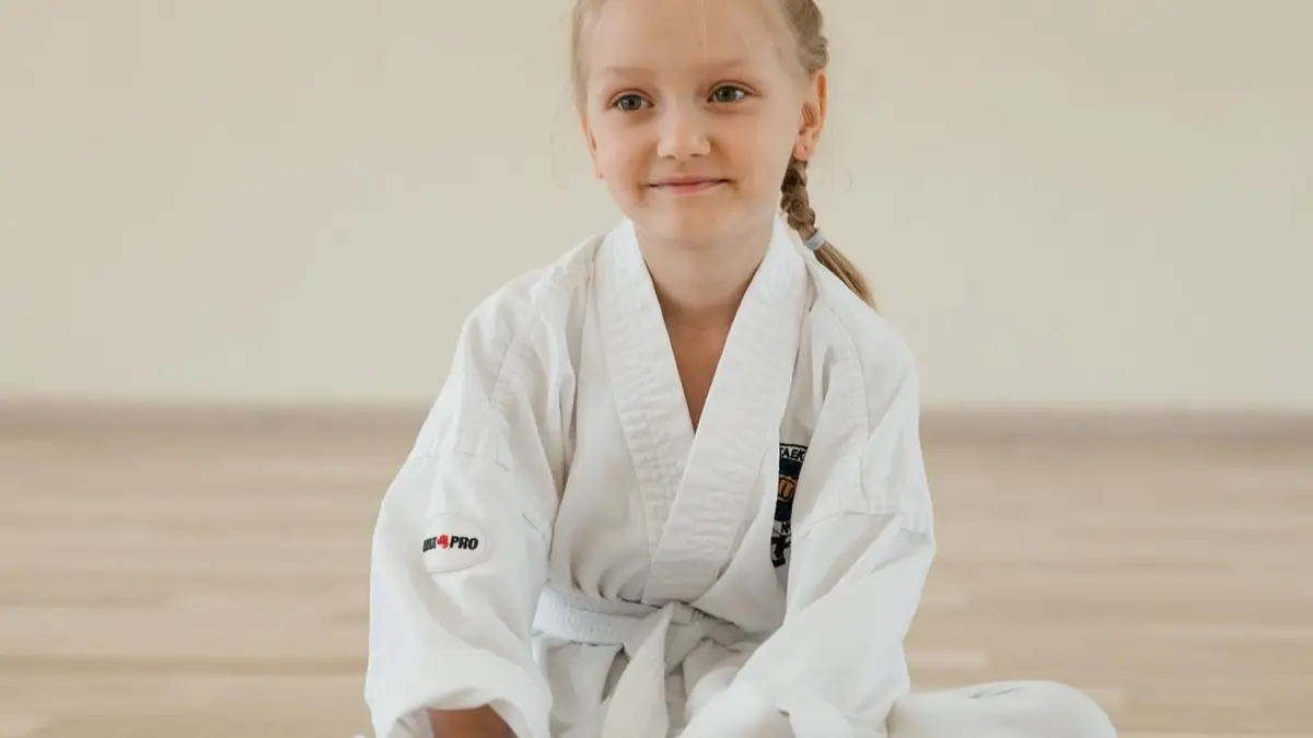 Девочка в добоке сидит на полу зала и держит в руках белый пояс