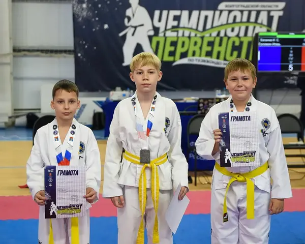 Трое мальчиков в добоках с медалями и грамотами на фоне баннера чемпионата и первенства Волгоградской области