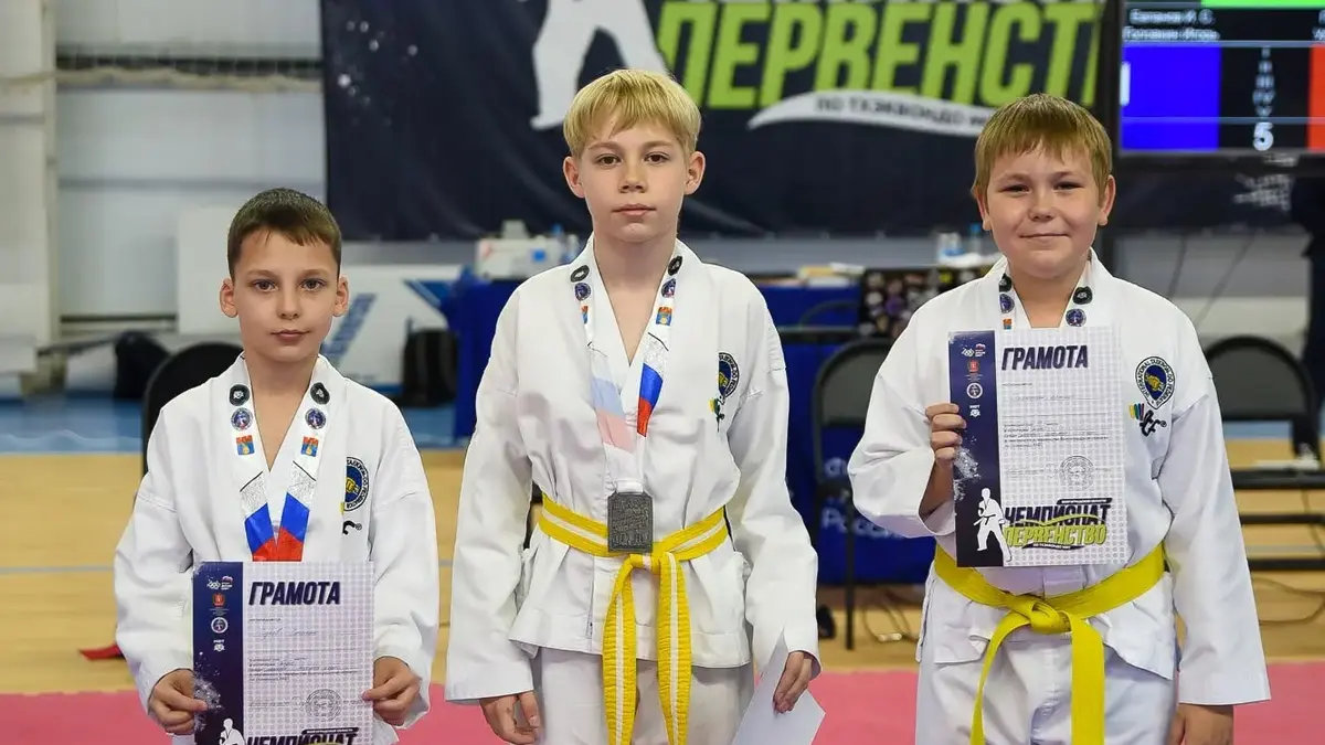 Трое мальчиков в добоках с медалями и грамотами на фоне баннера чемпионата и первенства Волгоградской области